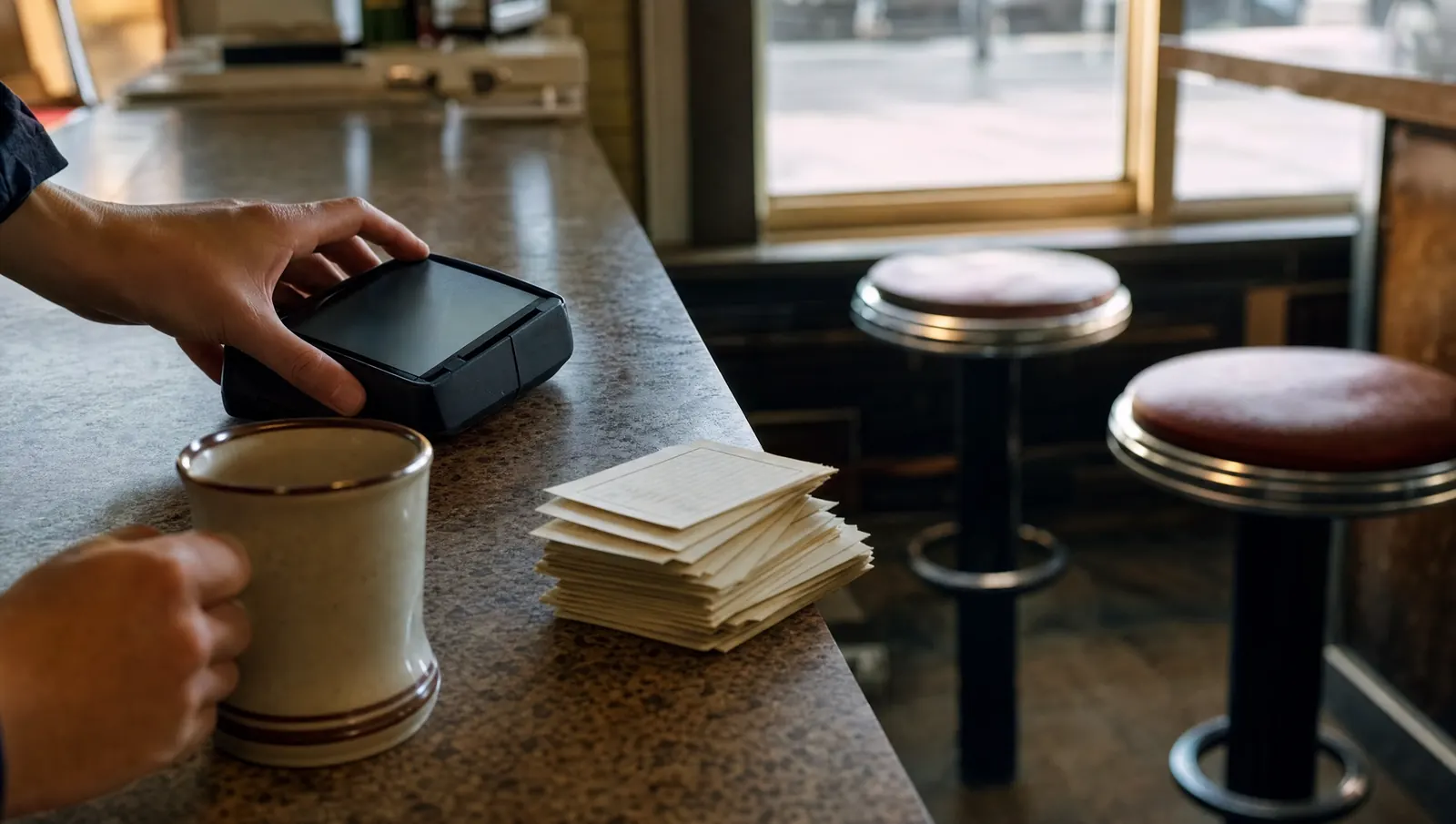 A small diner service counter with an unbranded countertop payment terminal beside a coffee mug and face-down order tickets.