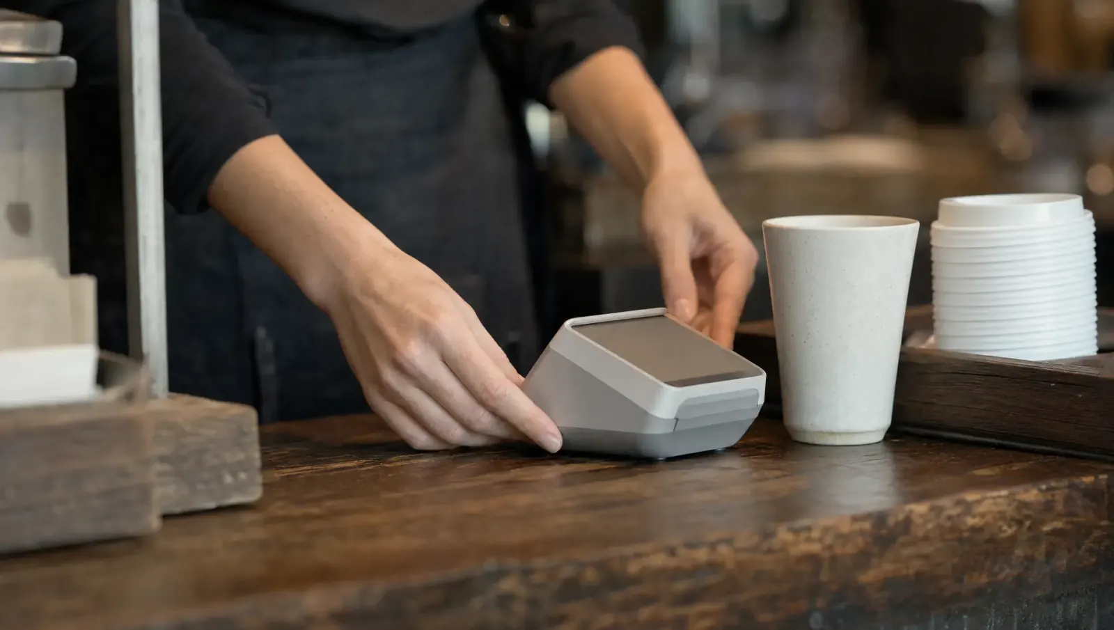 A barista's hands setting an unbranded countertop payment terminal on a wood coffee-shop counter beside a ceramic cup and a stack of paper cups.