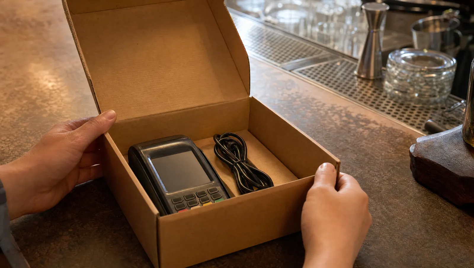 An unplugged plain countertop payment terminal sitting in an open cardboard box on a bar counter, cable coiled beside it.