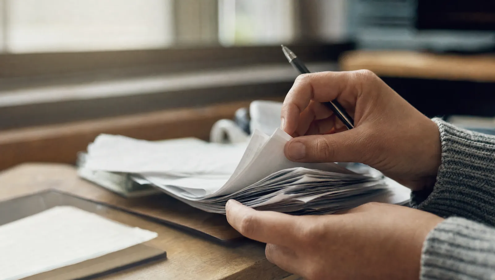 A pair of hands flipping through a stack of folded paper statements at a back desk, a pen tucked behind.